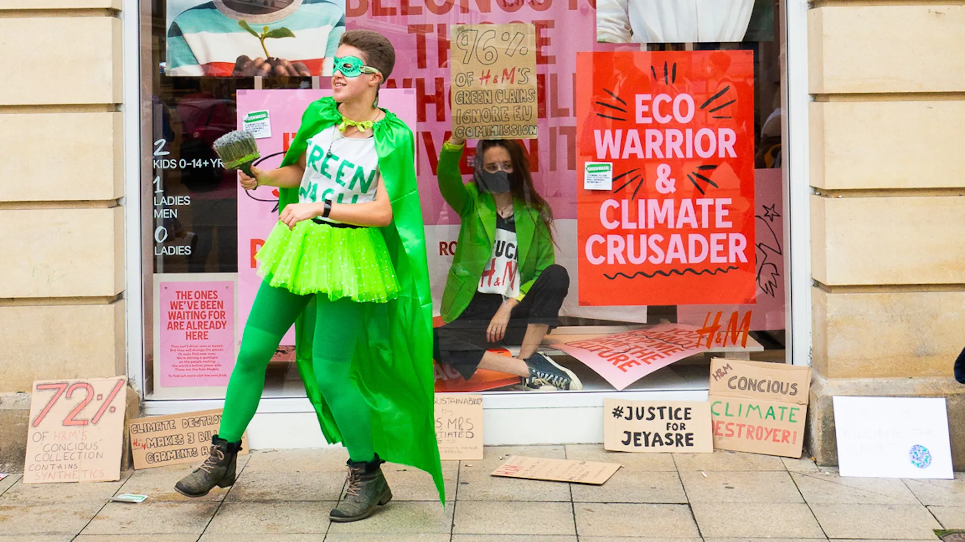 Deux militants vêtus de costumes verts de type « éco-héros » manifestent devant une vitrine de magasin H&M, entourés de pancartes dénonçant le greenwashing et la crise climatique.